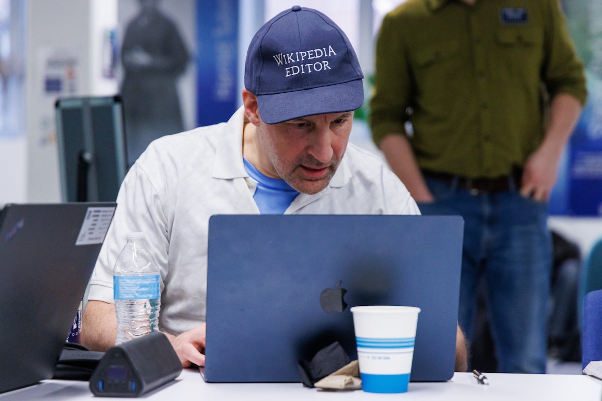 A man wearing a baseball cap that says "Wikipedia Editor." He is sitting at table typing on a laptop computer.
