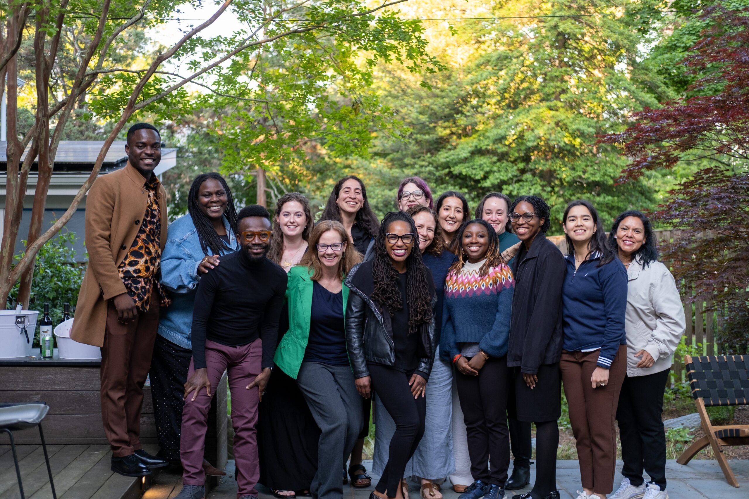 A group photo of 15 G+JI faculty, staff, and fellows standing outside and smiling. Behind them are lush green trees.