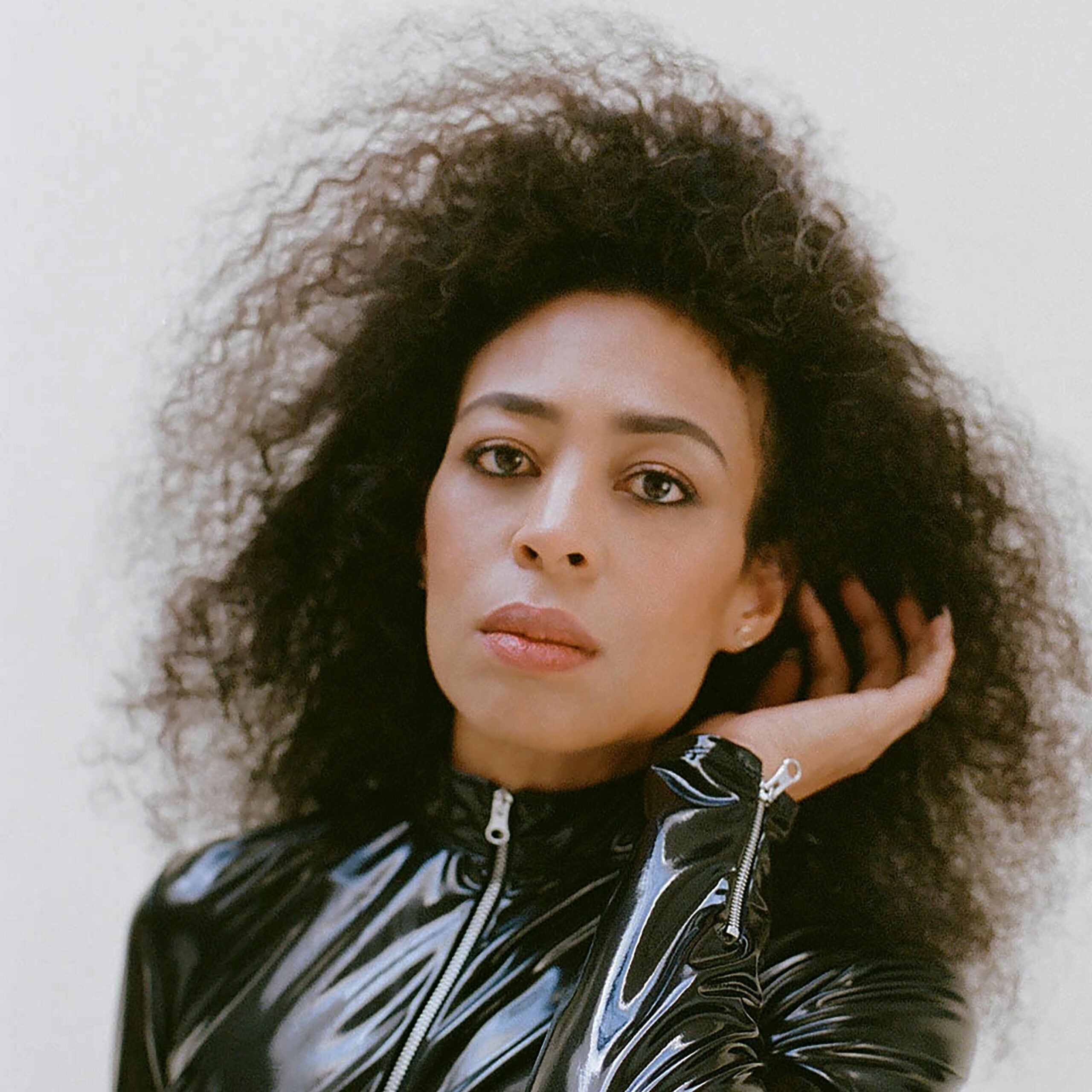 Portrait of Candice Hoyes wearing a glossy black top, with  curls against a light background.